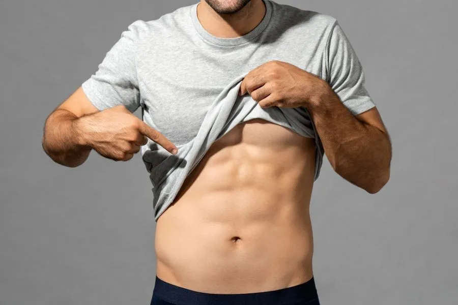 A man lifts his shirt and shows his abs while pointing at his stomach